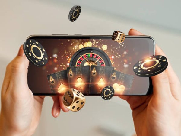 Creative background, online casino, in a man's hand a smartphone with playing cards, roulette and chips, black-gold background. Internet gambling concept. Copy space.